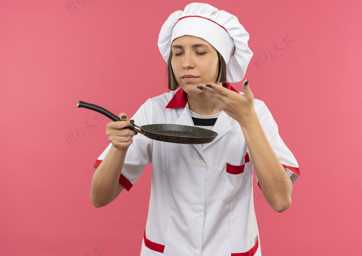 厨师身着厨师制服的年轻女厨师手拿煎锅 闭着眼睛闻着 手放在粉色背景上隔绝的空气中制服持有薯条