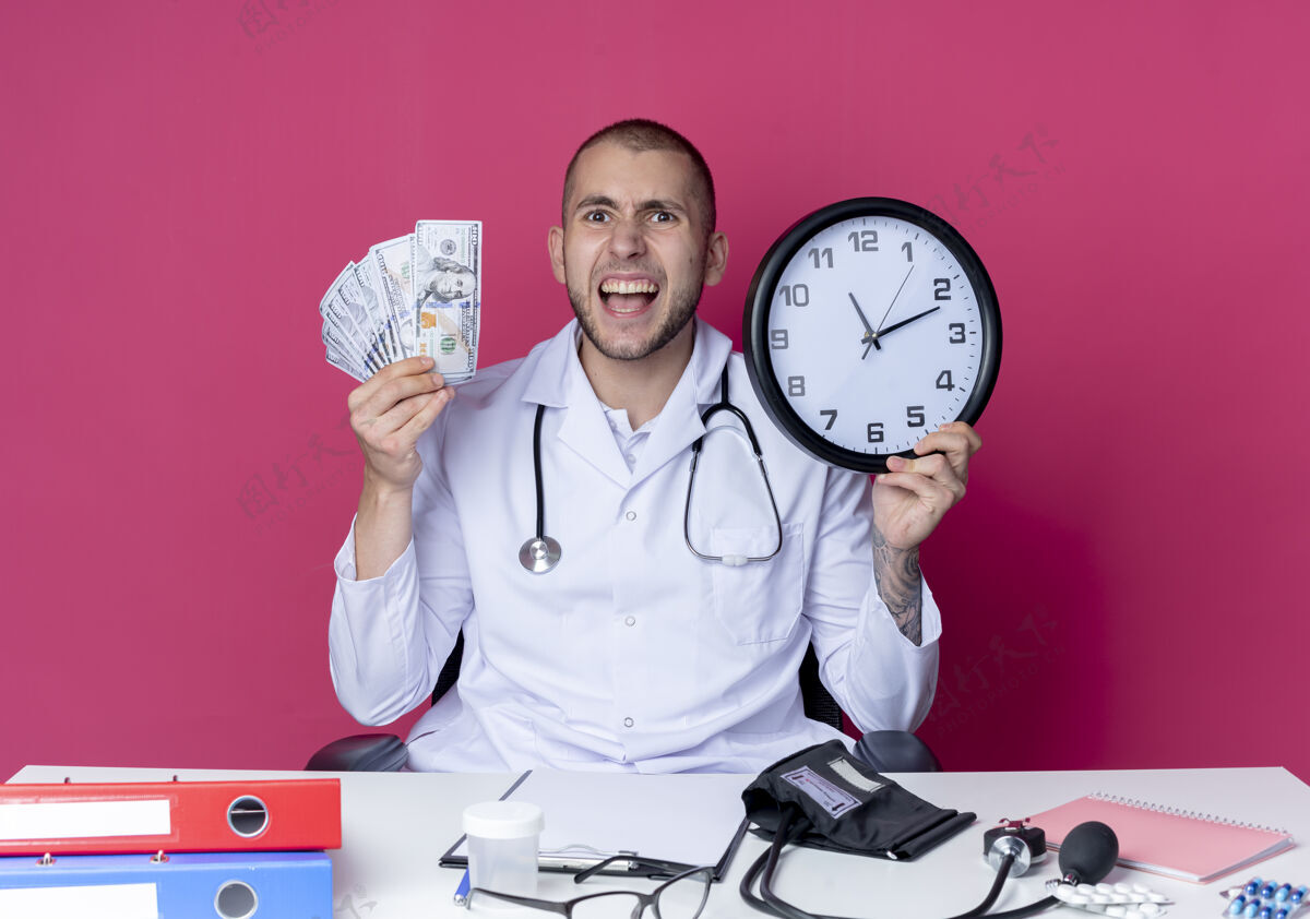 年轻年轻的男医生穿着医用长袍 听诊器坐在办公桌旁 手里拿着工作工具 手里拿着钱 粉色的钟被隔离开来办公桌医生长袍