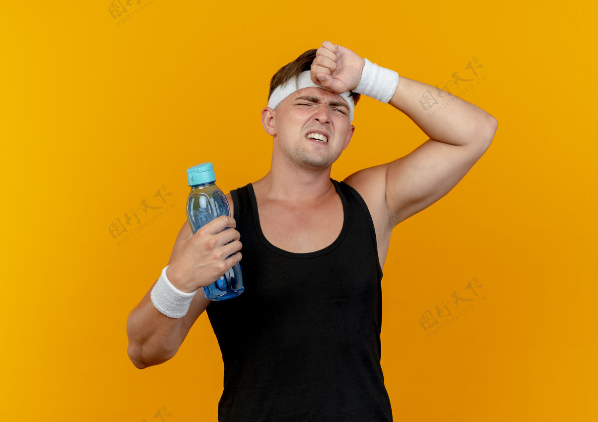 手疲惫的年轻帅气的运动型男人戴着头带和腕带拿着水瓶 把手放在孤立在橙色背景上的额头上头带瓶子抱着