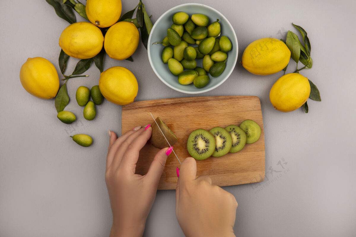 食物女性手在木制厨房板上切割猕猴桃的俯视图 刀子上放着金盏花 碗里的柠檬被隔离在白色背景上水果手顶部