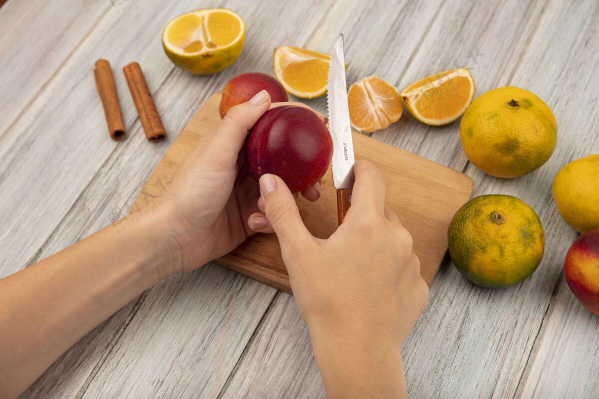 饮食俯视图中的女性双手在木制厨房板上用刀切桃子 橘子被隔离在灰色的木制背景上顶部新鲜板