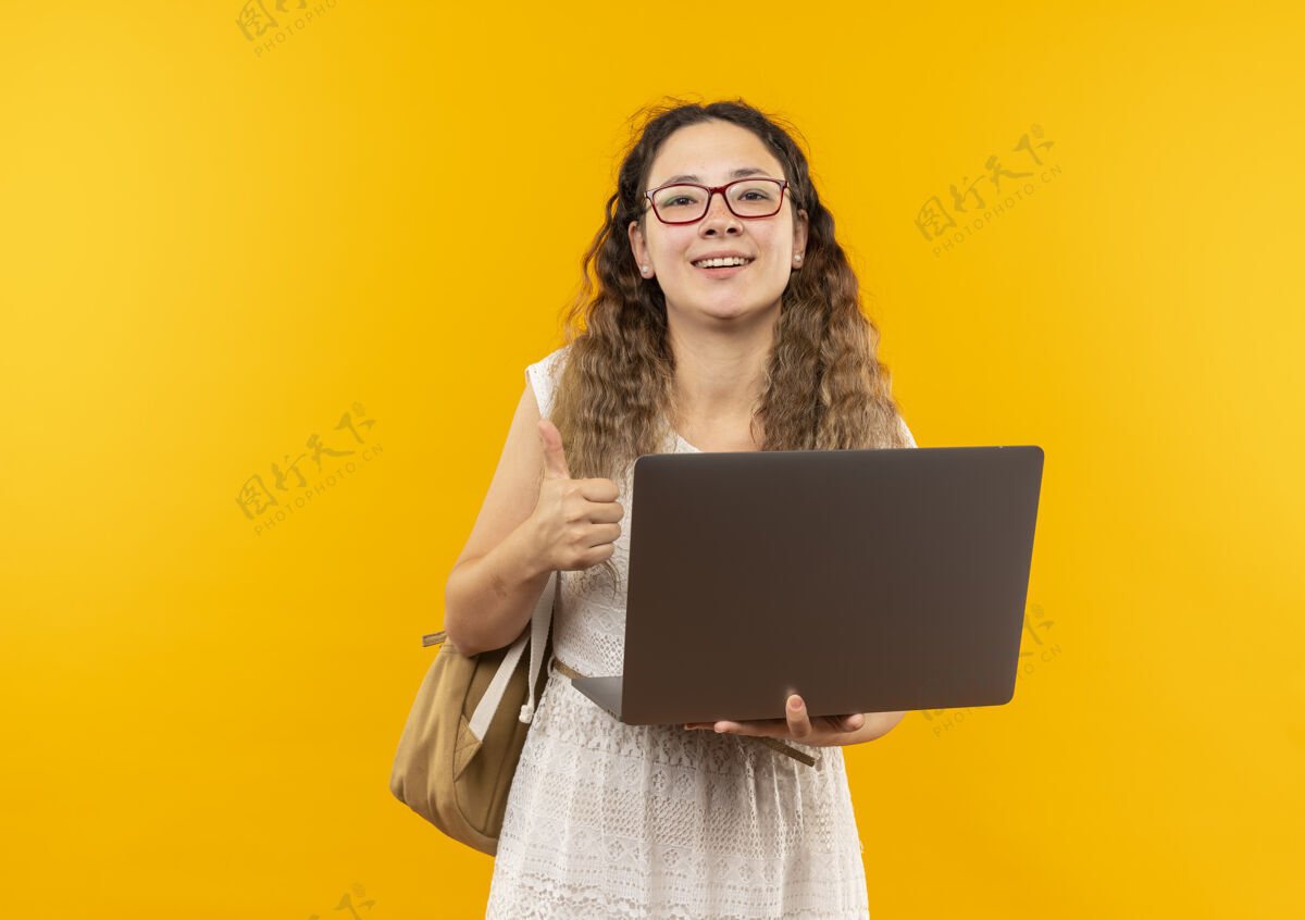 黄色快乐的年轻漂亮的女学生戴着眼镜 背着手提电脑 在黄色的地板上竖起大拇指背部表演快乐