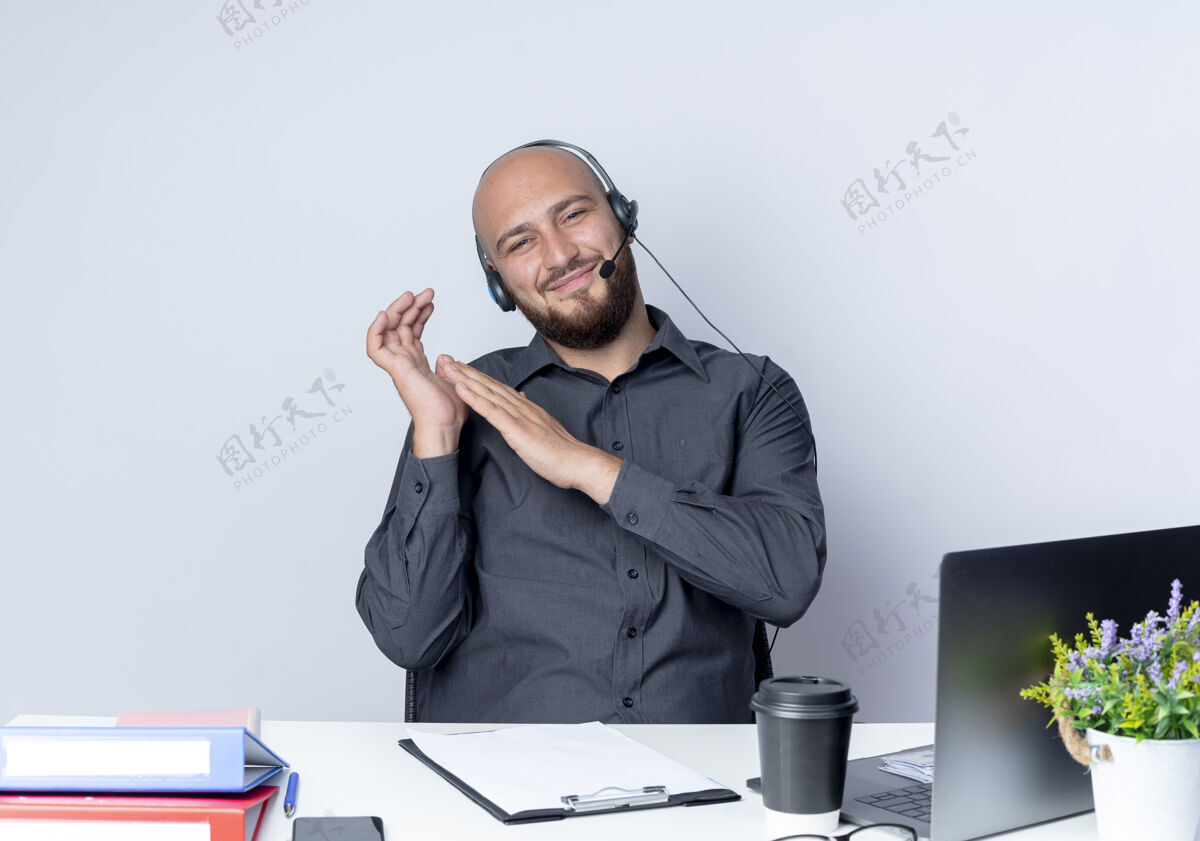 请高兴的年轻秃头呼叫中心男子戴着耳机坐在办公桌旁与工作工具保持手靠近另一个孤立的白色背景工作戴着工具