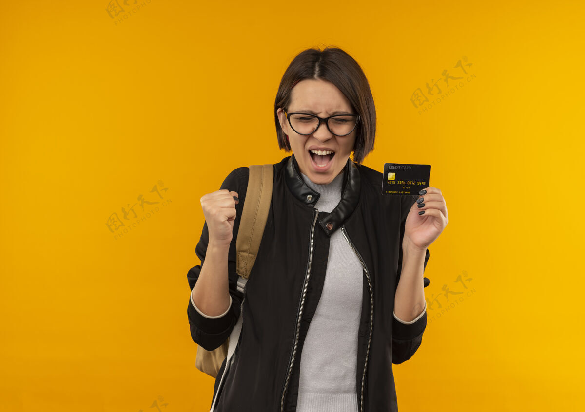 关闭快乐的年轻学生女孩戴着眼镜 背着包拿着信用卡紧握拳头闭着眼睛孤立在橙色信用包喜悦