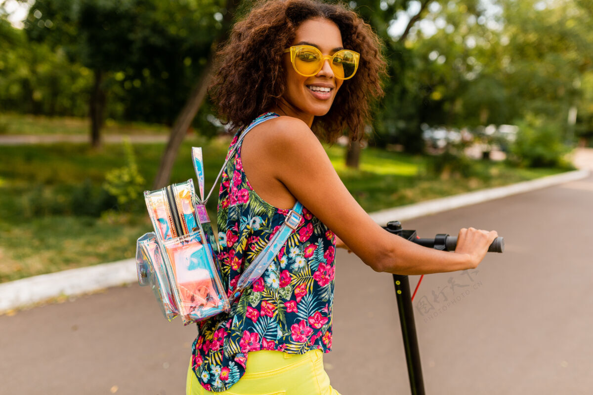 女性年轻时尚的黑人女子在公园里玩得很开心骑着电动脚踏车在夏天的时尚风格 五颜六色的时髦服装 穿着背包和黄色太阳镜环保积极车辆
