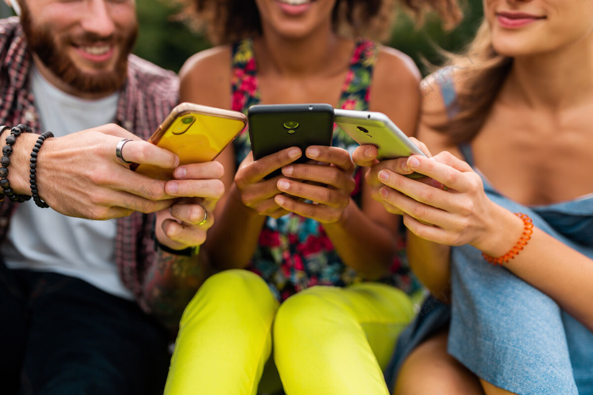 手机快乐的年轻朋友们坐在公园里用智能手机微笑 男人和女人一起玩应用程序多种族多民族