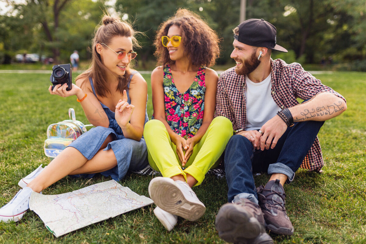 团体快乐的年轻人伴着谈笑风生的朋友坐在公园里 男女同乐 缤纷的夏日潮人时尚风格 带着相机旅行微笑太阳镜朋友
