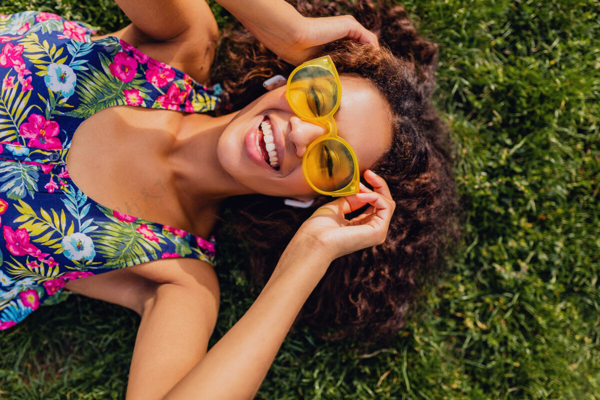 女性年轻时尚的黑人女子戴着无线耳机听音乐在公园里玩得很开心 夏日时尚风格 五颜六色的时髦装扮 躺在草地上 俯瞰风景年轻人物非洲裔美国人