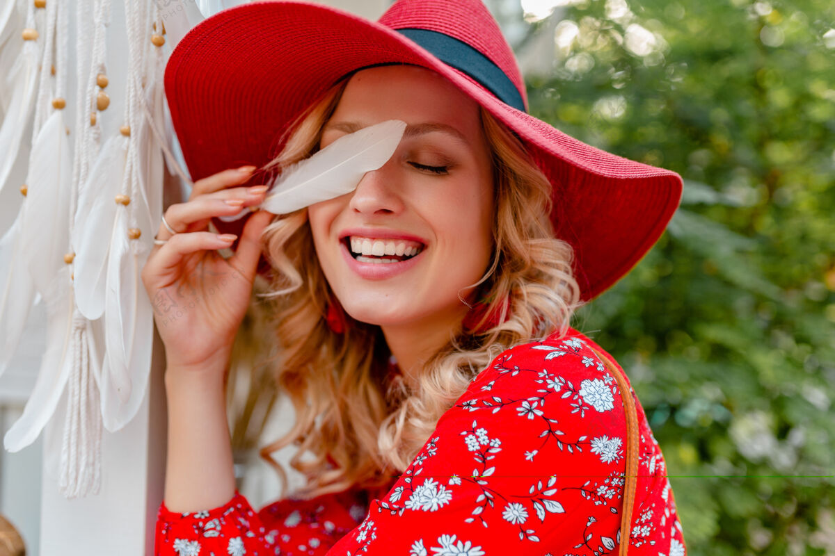 女性近距离肖像迷人时尚金发微笑的妇女在稻草红色帽子和衬衫夏季时尚服装举行白色羽毛性感性感的面部皮肤休闲装束配饰
