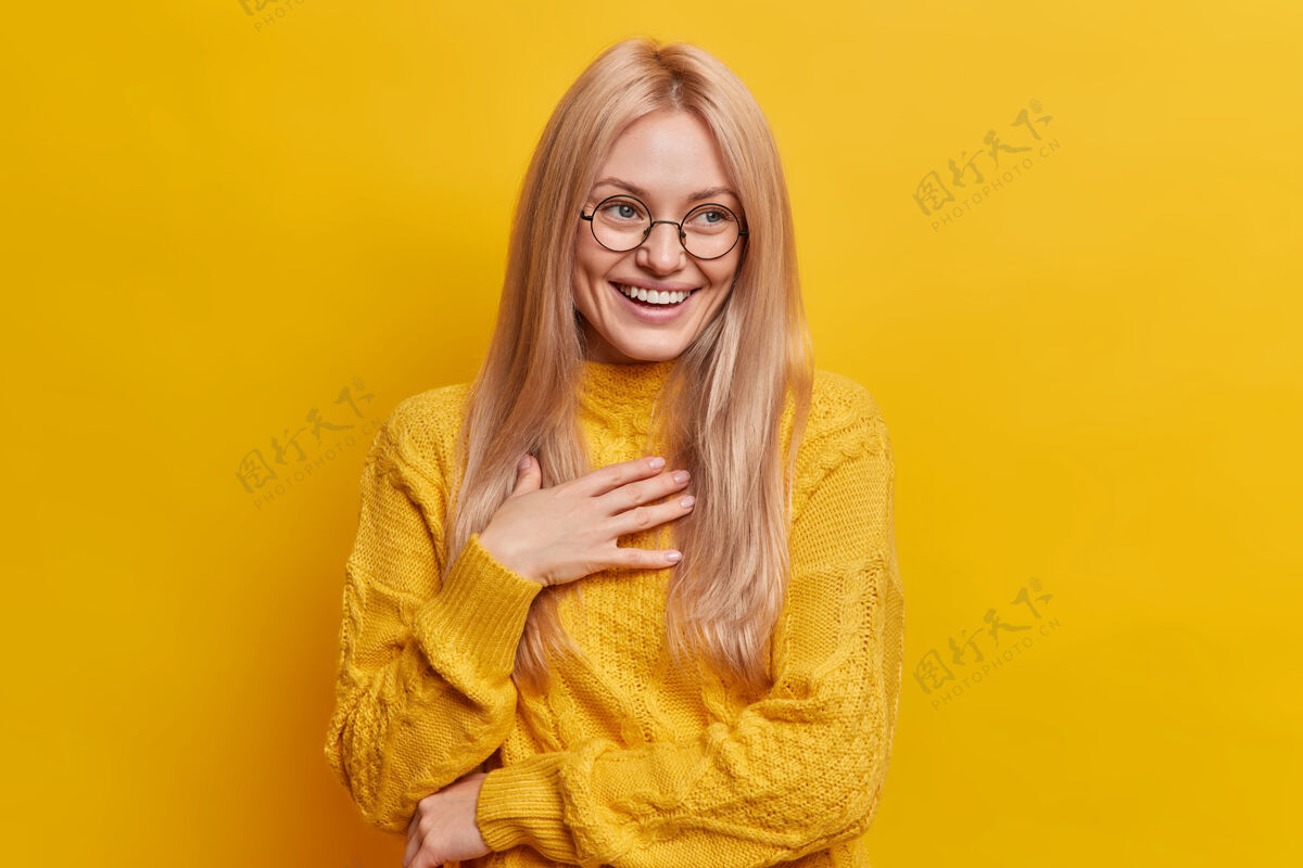 单色快乐的金发欧洲女人开心地笑看起来无忧无虑的咯咯笑积极地把手放在胸前眼镜欢呼情绪