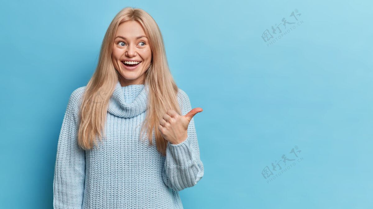 姿势快乐的金发欧洲女人穿着针织毛衣有着愉快的表情 拇指指向复制空间微笑手指肖像