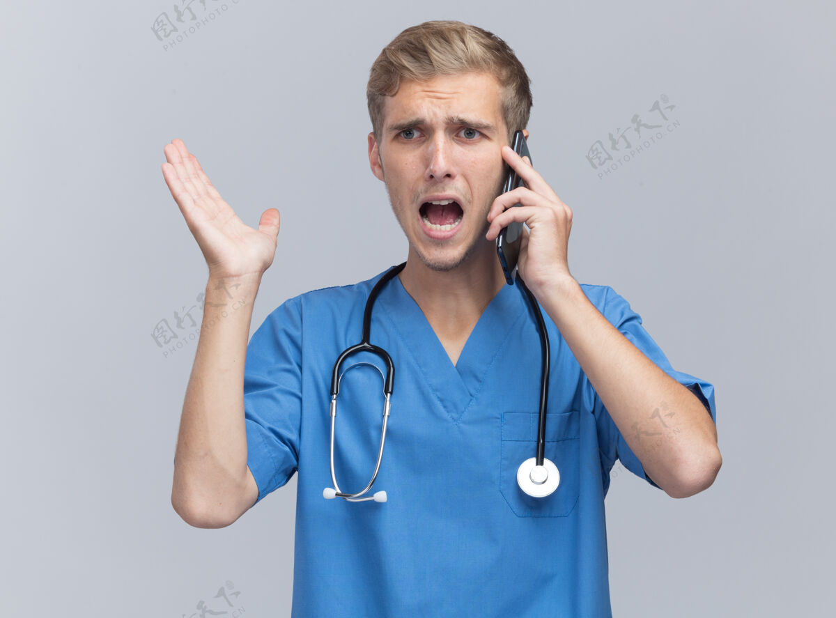 电话愤怒的年轻男医生穿着医生制服 手持听诊器在电话里讲话 手被隔离在白墙上制服愤怒传播