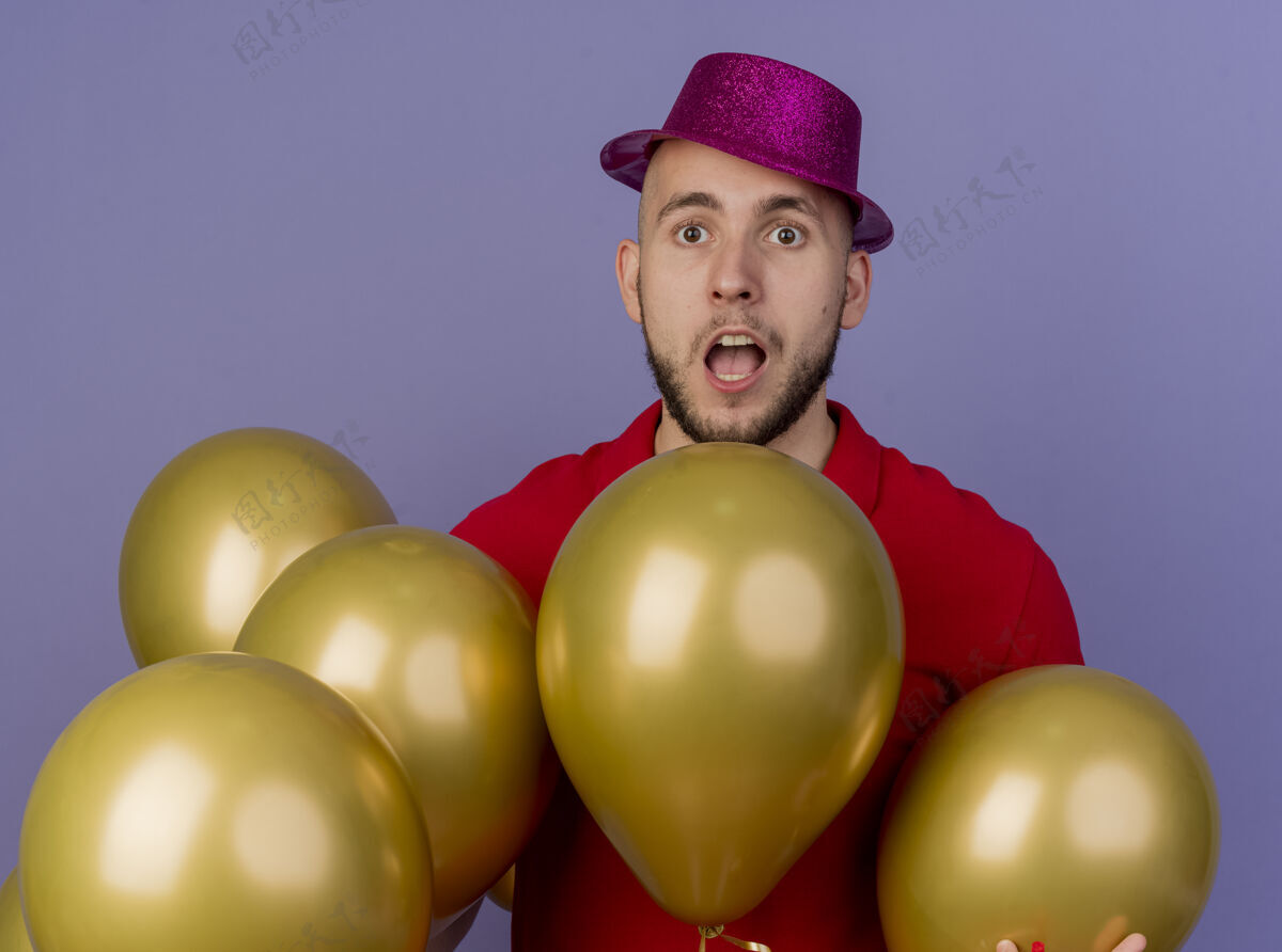 站着令人印象深刻的年轻英俊的斯拉夫党人戴着党的帽子站在气球后面看着紫色背景上孤立的相机紫色年轻人印象