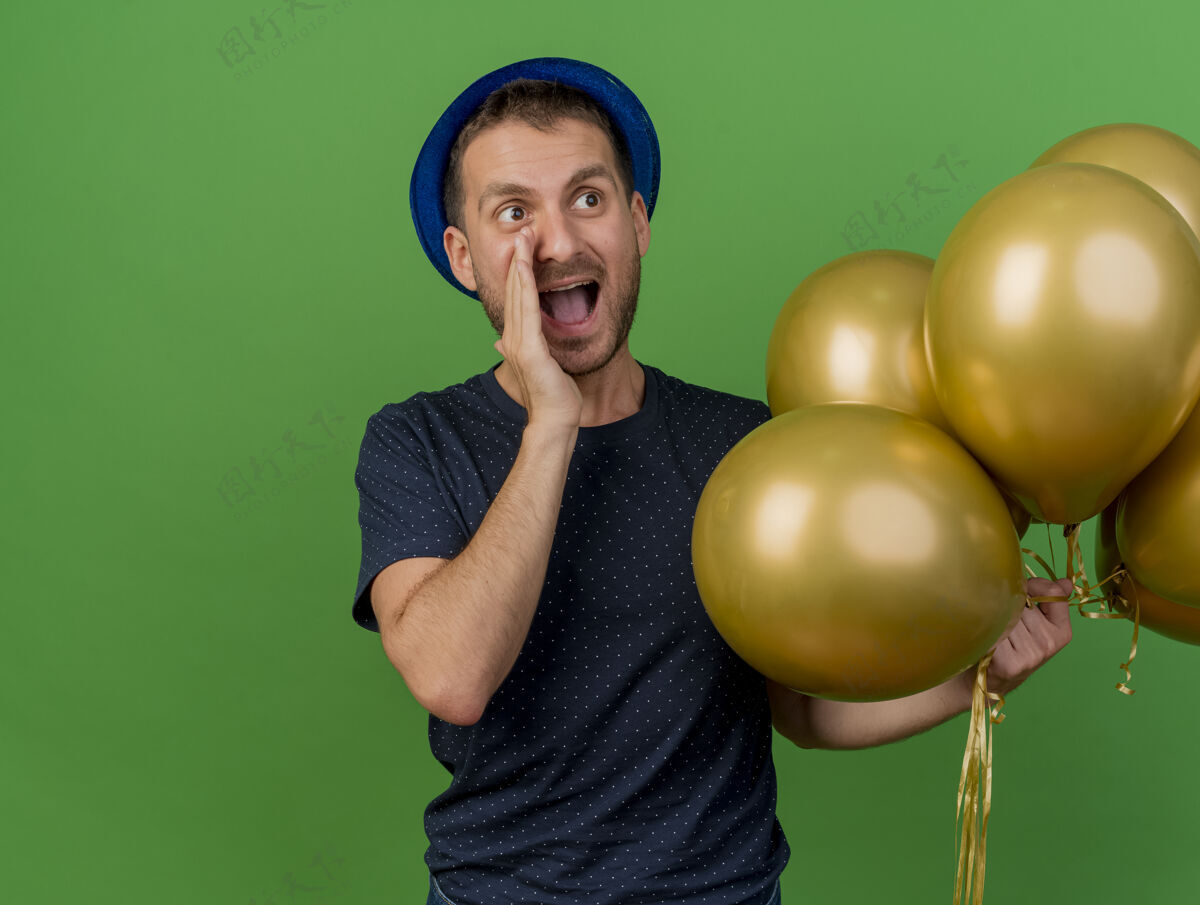 聚会一个高加索帅哥 戴着蓝色的派对帽 手里拿着氦气球 一边看着一边喊着一个被隔离在绿色背景下的人举行男人兴奋