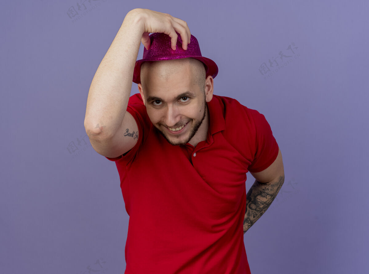 斯拉夫面带微笑的年轻英俊的斯拉夫党人戴着党的帽子抓住它看着相机保持手背后紫色背景上的复制空间隔离男人空间紫色