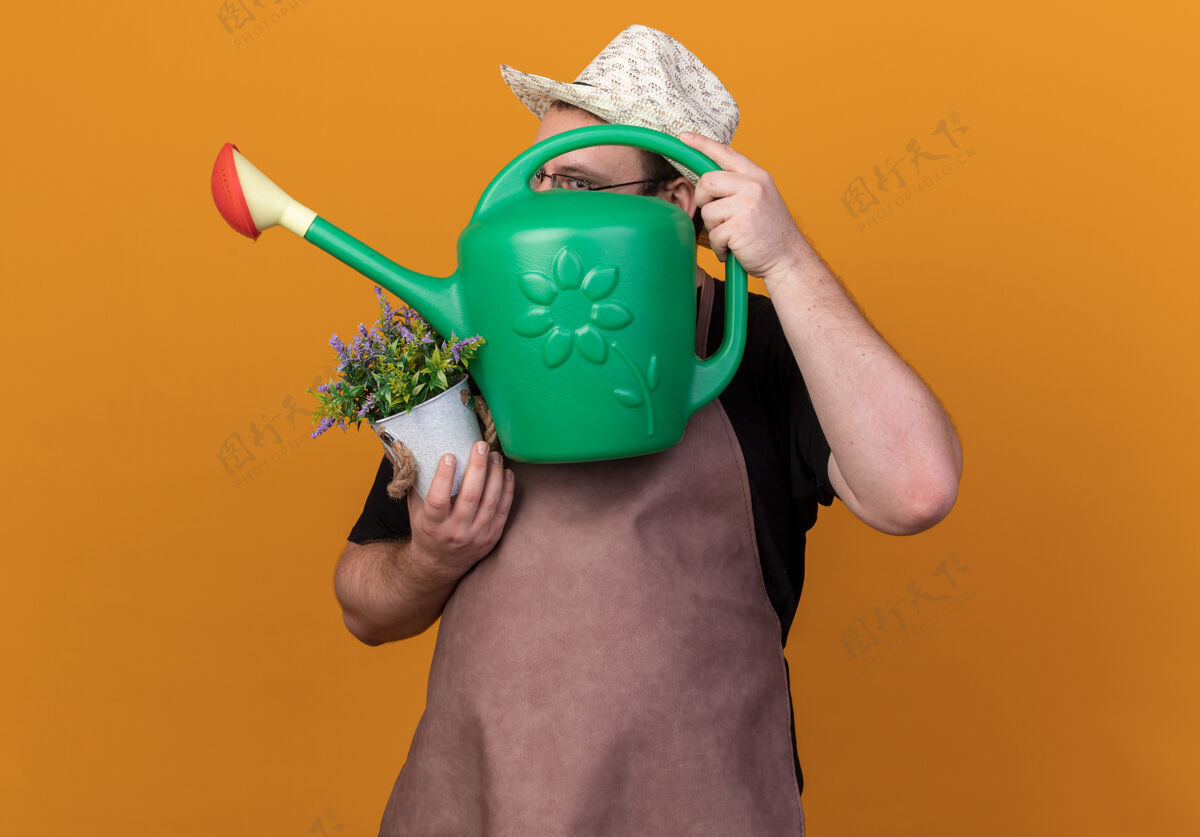 喜悦快乐的年轻男园丁戴着园艺帽和手套 脸上蒙着浇水罐 手里拿着隔离在橙色墙上的花盆里的花男穿园艺