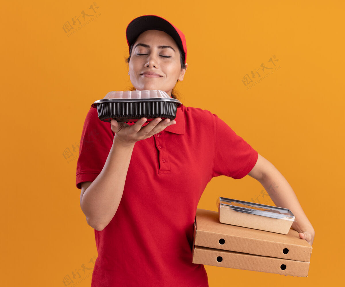 请年轻的送货员穿着制服 戴着帽子 手里拿着披萨盒 嗅着食物 被隔离在橙色的墙上穿披萨帽子