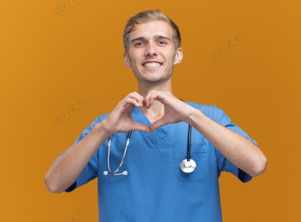 手势微笑着的年轻男医生穿着医生制服 带着听诊器 在橙色的墙上显示着孤立的心脏姿势男性年轻制服