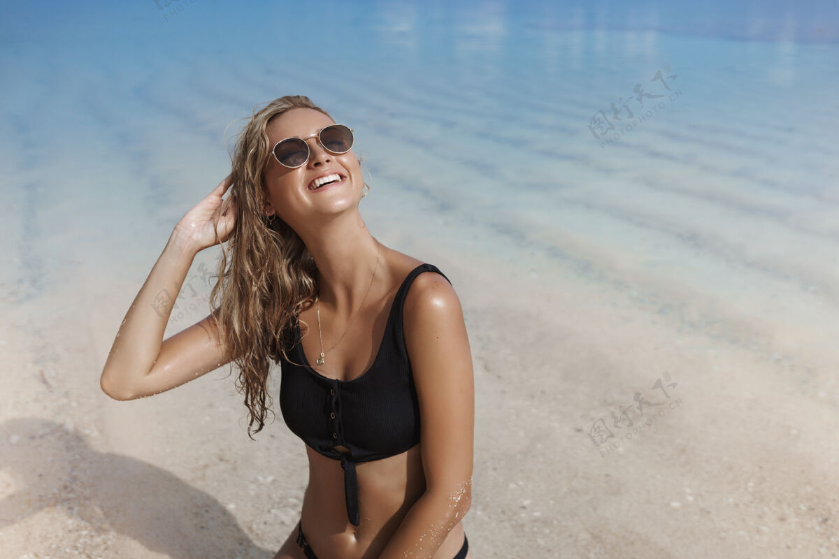 海岸戴着墨镜和比基尼的金发女人抬起头 高兴地微笑着放松娱乐比基尼