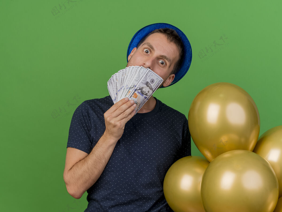 空间一个戴着蓝色派对帽的高加索帅哥 手里拿着氦气球和钞票 背景是绿色的 上面留着复制空间气球聚会人