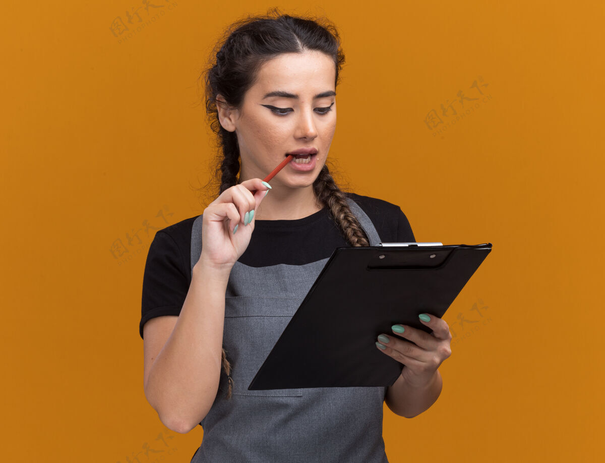 年轻高兴的年轻女理发师拿着制服 看着剪贴簿把铅笔放在隔离在橙色墙上的嘴上剪贴板女理发师
