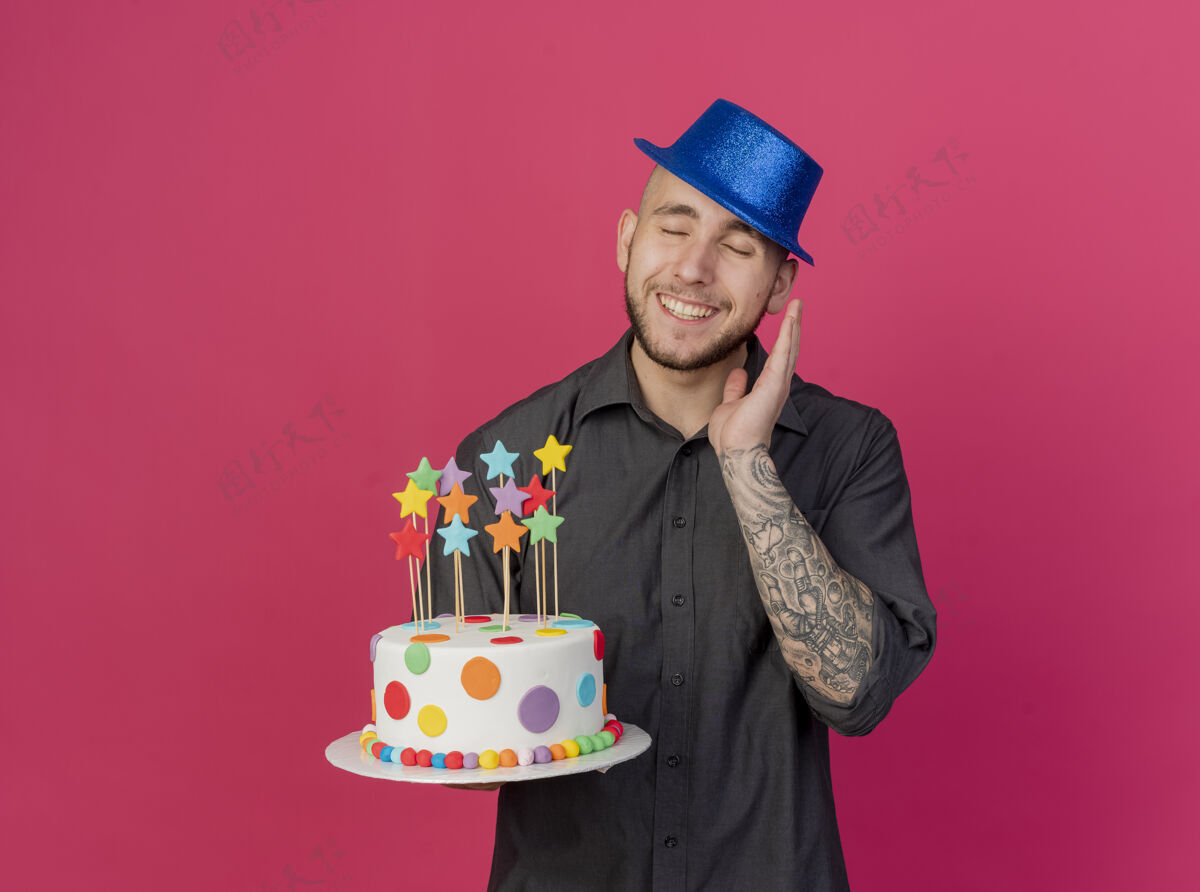 生日微笑的年轻帅哥斯拉夫党人戴着党的帽子拿着生日蛋糕与明星保持手在空气中闭上眼睛在深红色背景与复制空间隔离蛋糕帽子年轻