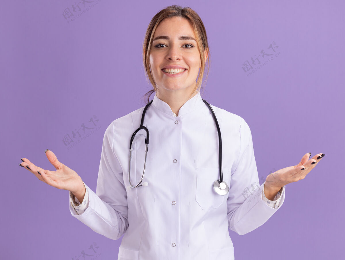 医疗面带微笑的年轻女医生穿着医用长袍 听诊器摊开双手隔离在紫色的墙上传播长袍手
