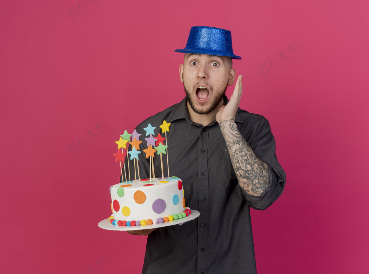 手惊讶的年轻英俊的斯拉夫党人戴着党的帽子拿着生日蛋糕与明星保持手在空中看着摄像机在深红色背景与复制空间隔离深红色小伙子生日