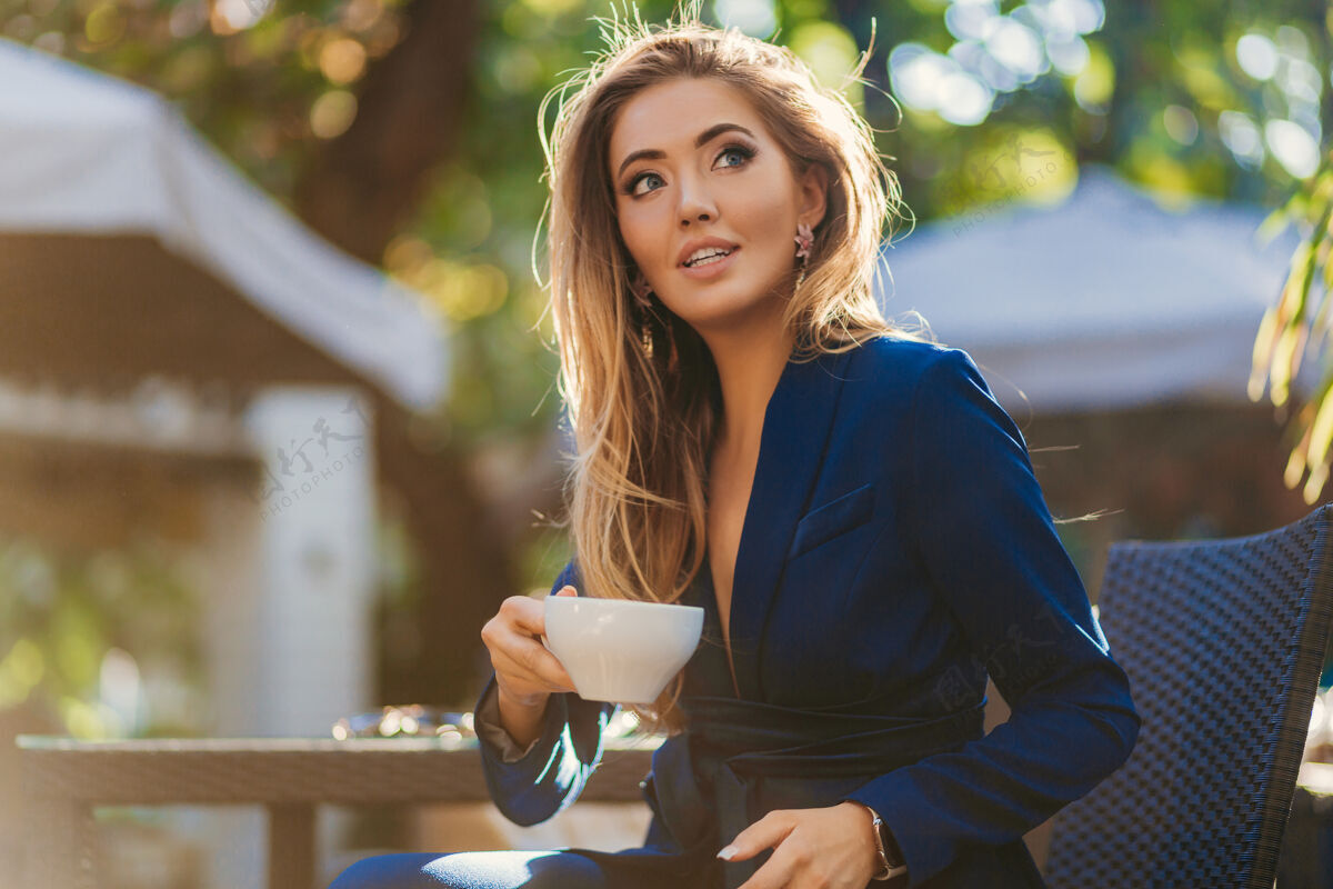 街头风格穿着蓝色优雅套装的迷人时尚女士坐在咖啡馆的桌子旁喝着一杯卡布奇诺优雅现代魅力