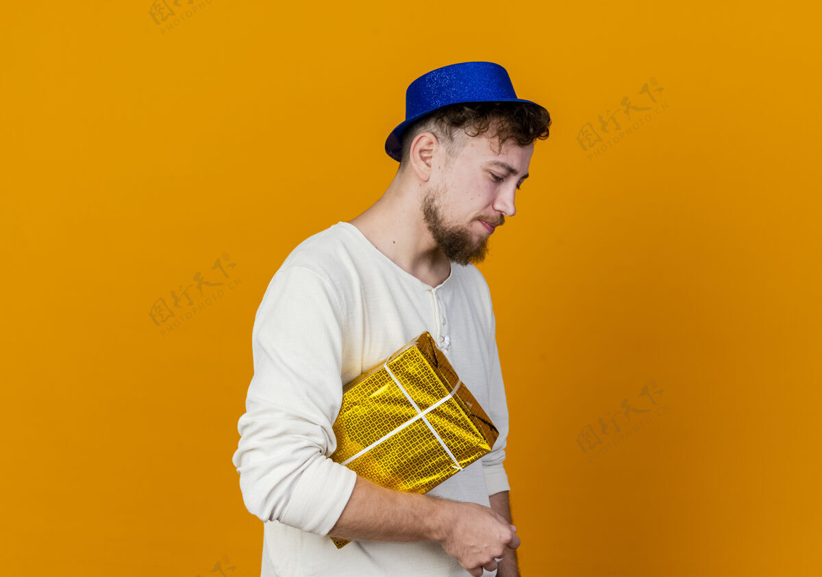 个人资料悲伤的年轻英俊的斯拉夫党人戴着党帽站在侧视图举行礼品盒向下看橙色背景与复制空间孤立站下空间