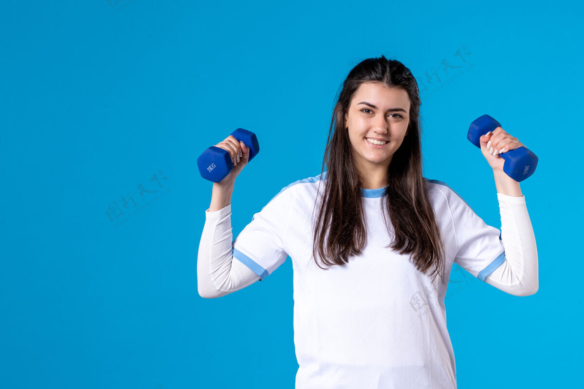 哑铃正面图：年轻女子手持蓝色哑铃站在蓝色墙上锻炼体重赢家