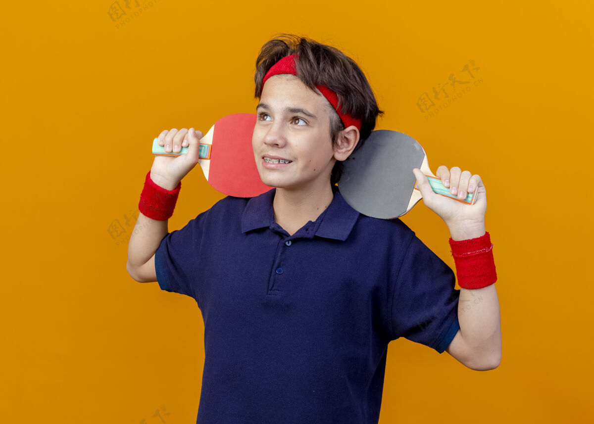 球拍面带微笑的年轻帅气的运动男孩戴着头带和护腕 戴着牙套 肩上扛着乒乓球拍 看着橙色背景下孤立的一面运动微笑乒乓球