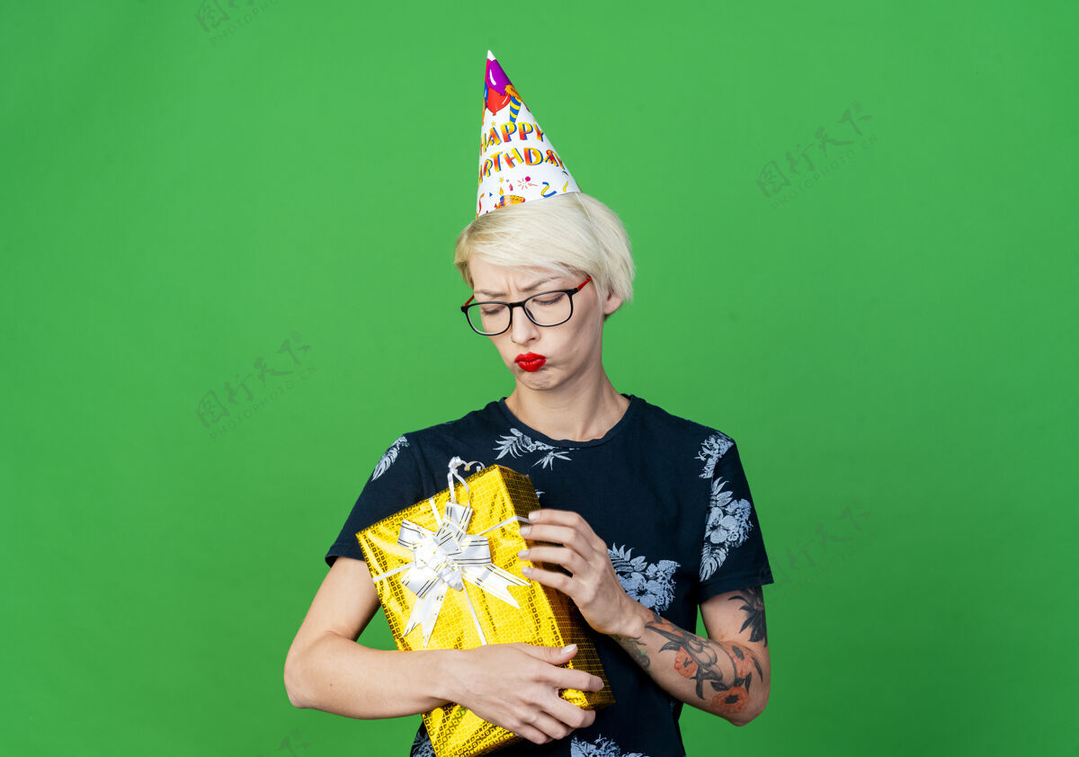 空间年轻的金发派对女孩戴着眼镜 戴着生日帽 拿着礼物盒 看着绿色的背景和复制空间隔离礼物聚会年轻