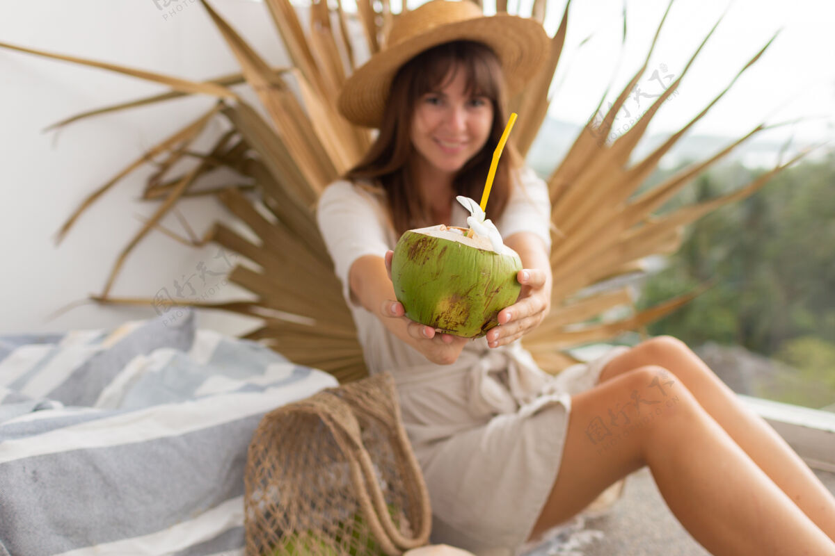 草帽戴着草帽 穿着亚麻裙的黑发美女站在露台上 手里拿着新鲜的椰子成人美丽的人夏天