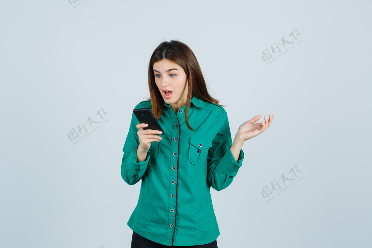时尚年轻女孩看着电话 惊讶地伸出手 穿着绿色上衣 黑色裤子 看上去很震惊 前视图嘴巴衬衫惊喜