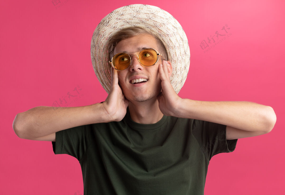 印象令人印象深刻的看边年轻帅哥穿着绿色衬衫 戴着眼镜戴着帽子把手放在脸颊上隔离在粉红色的墙上手衬衫壁板