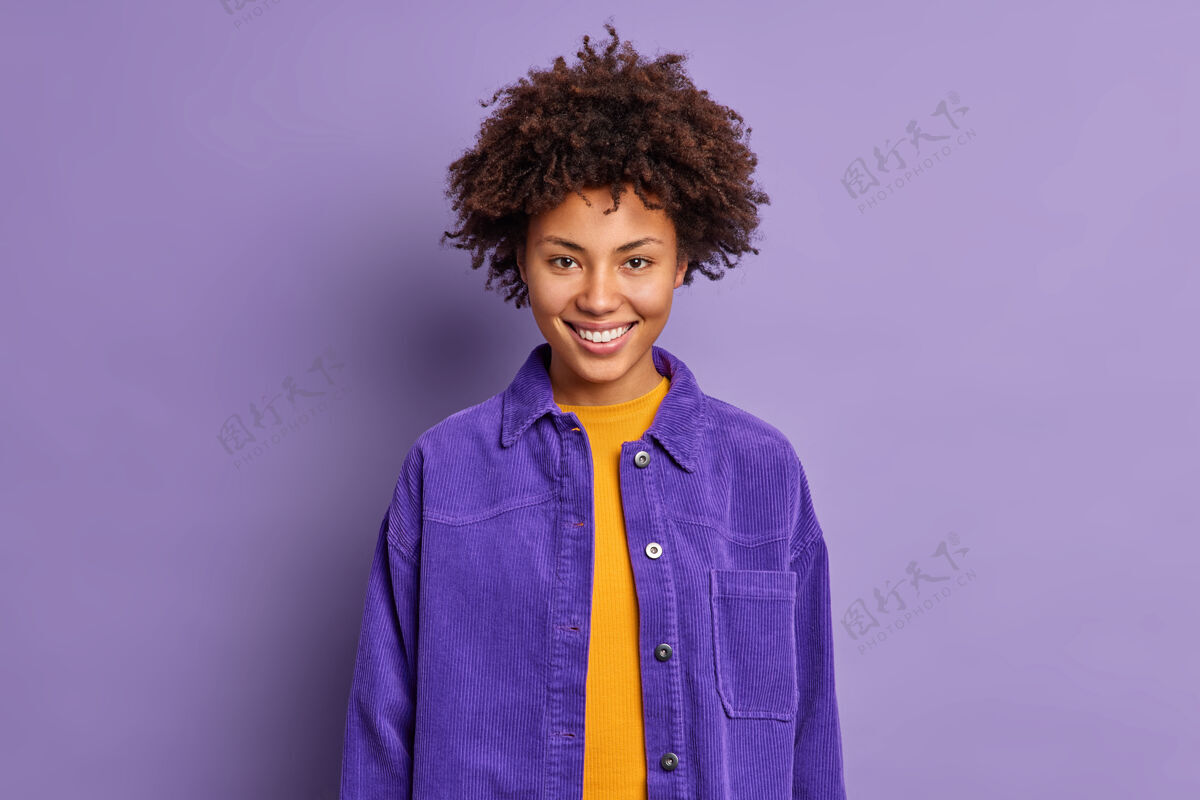 头发卷发美女的肖像笑容可掬地穿着时尚的紫色夹克 同一色调背景 心情愉悦地摆出愉快的姿势 与同事在室内交谈积极的情感理念女人种族满意