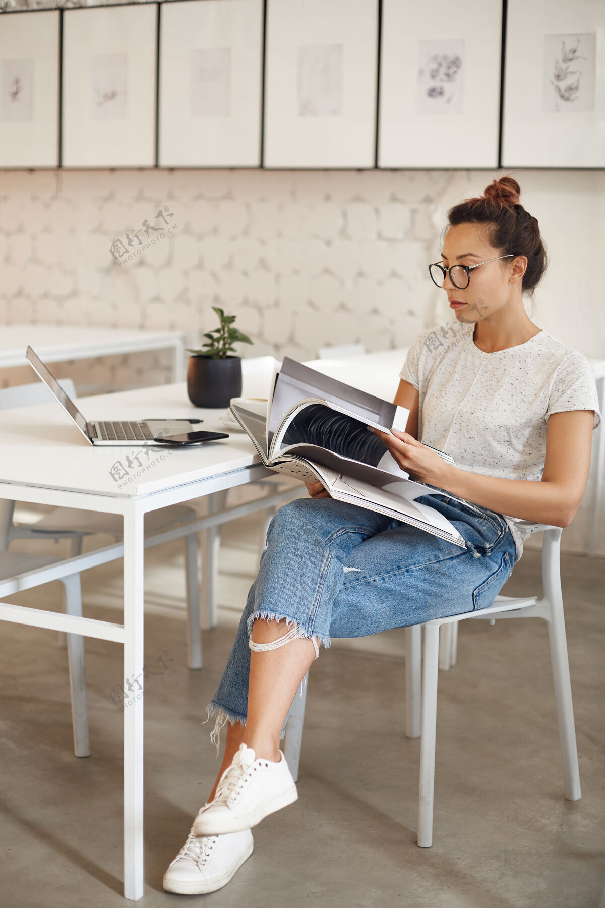 站立一位二十多岁的女士正在追求时尚设计的职业生涯 她坐在明亮的工作室里看着一本杂志 手里拿着笔记本电脑创意电脑在线