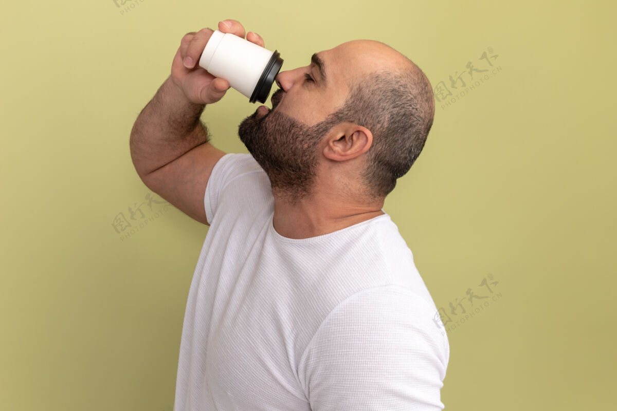 伙计站在绿色的墙上 一个留着胡子的男人穿着白色t恤 手里拿着咖啡杯要喝胡子咖啡杯子