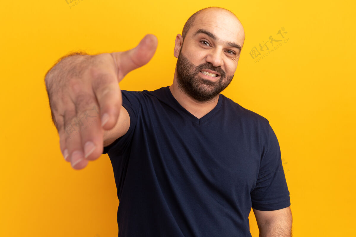 朋友留着胡子的男士身着海军蓝t恤 微笑友好地站在橙色的墙上向您致意立场手势提议