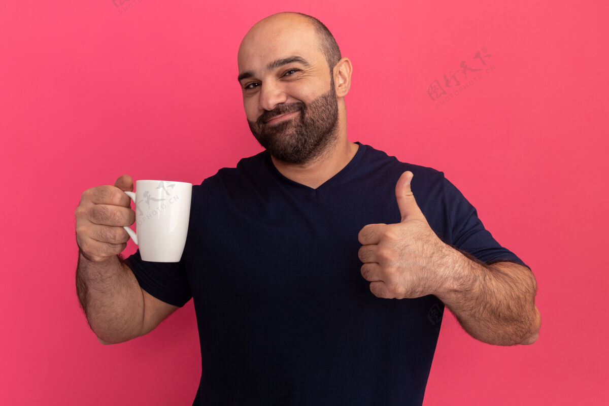 表演一个留着大胡子的男人穿着海军蓝t恤 手里拿着一个杯子 高兴而积极地竖起大拇指站在粉色的墙上男人正面杯子