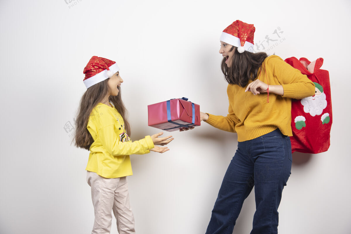女人送礼物给戴着圣诞老人红帽子的年轻女孩的女人女士年轻人年轻人