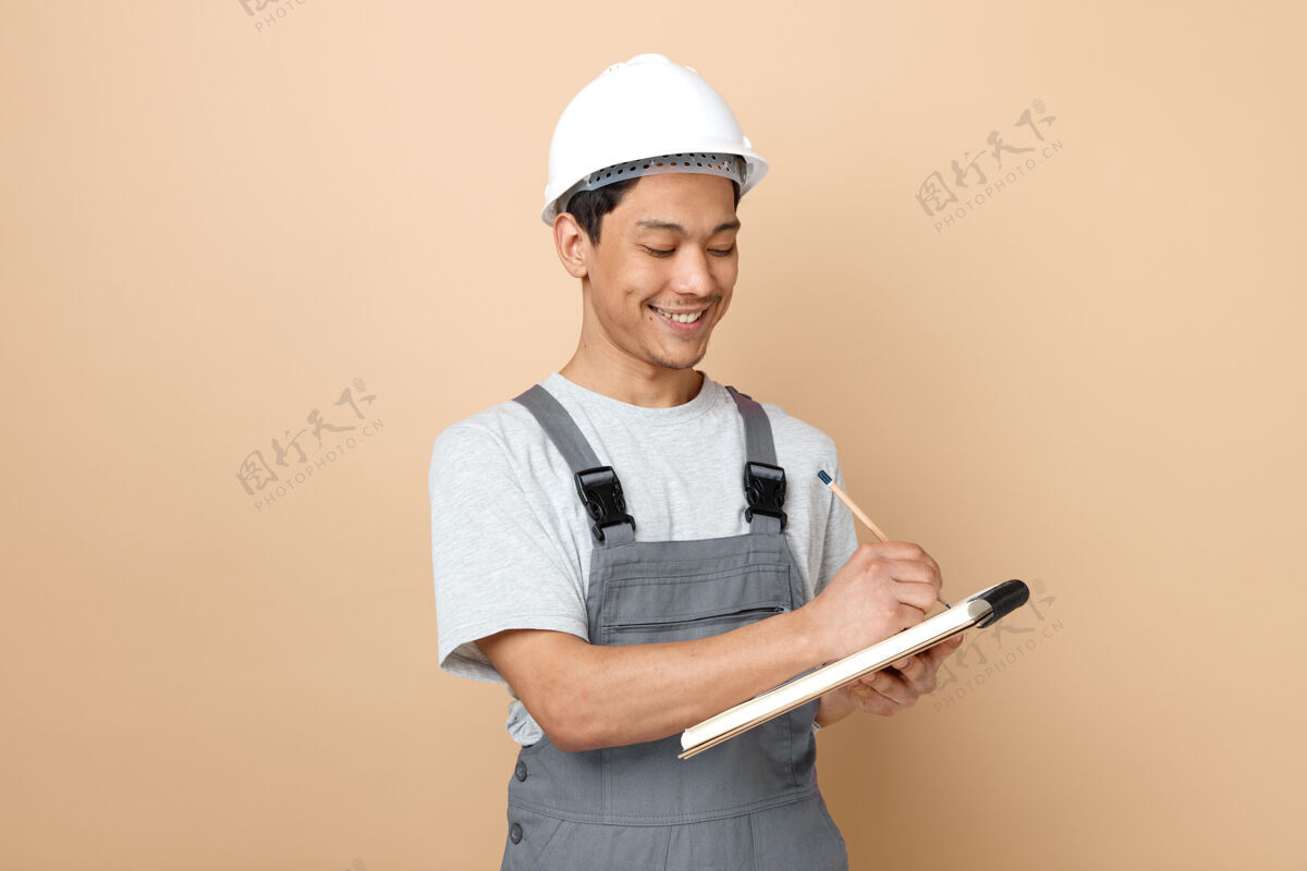 穿微笑的年轻建筑工人戴着安全帽 用铅笔在记事本上写着制服写作头盔工人