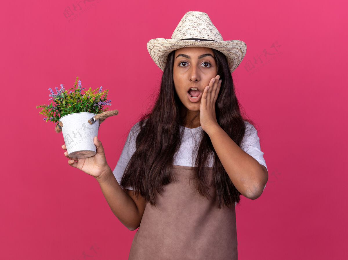 惊喜穿着围裙 戴着夏帽 手里拿着盆栽植物的年轻园丁女孩站在粉色的墙上 惊讶不已花园立场围裙