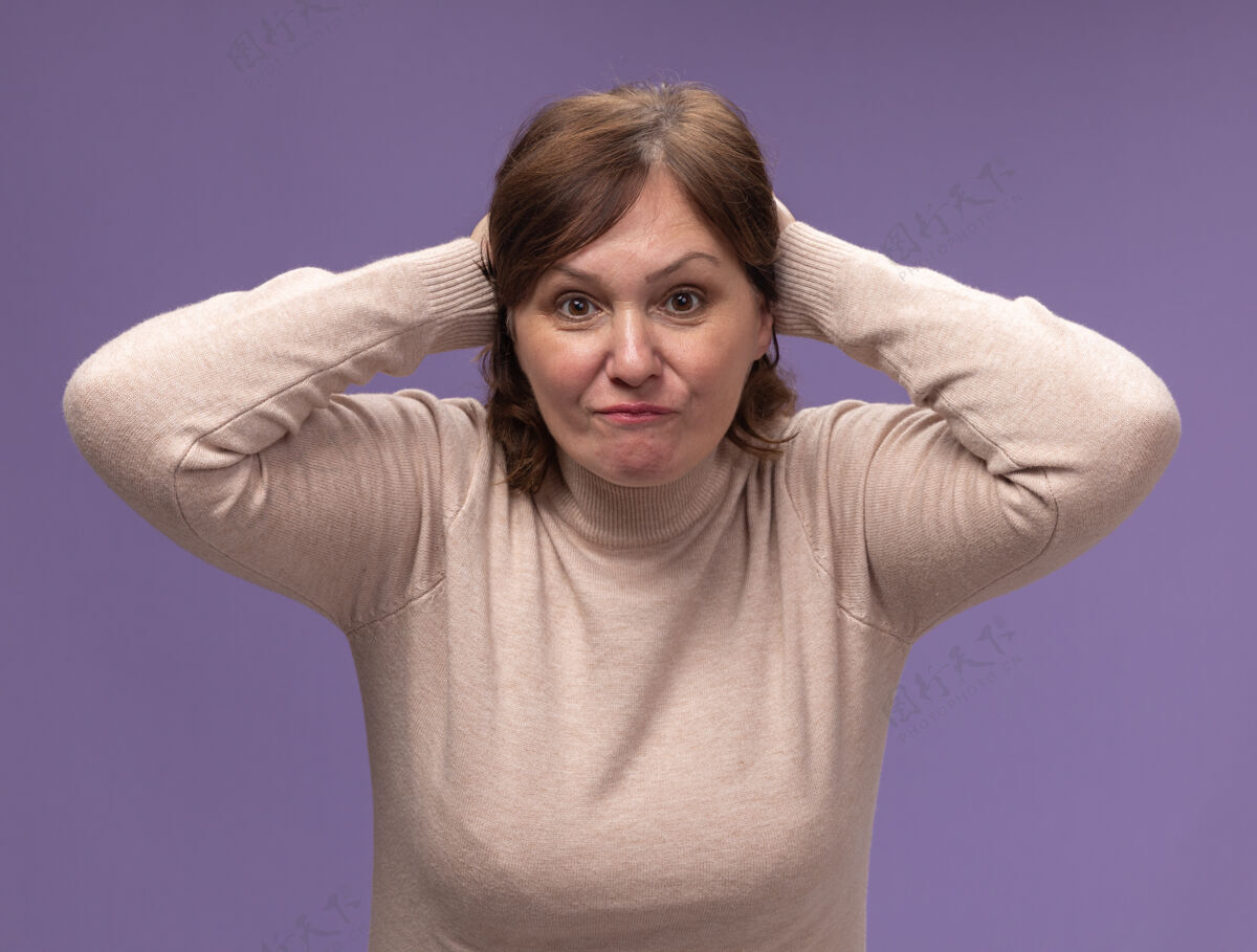 年龄穿米色高领套头衫的中年妇女站在紫色的墙上 双手放在头上 感到困惑和不快女人中等困惑