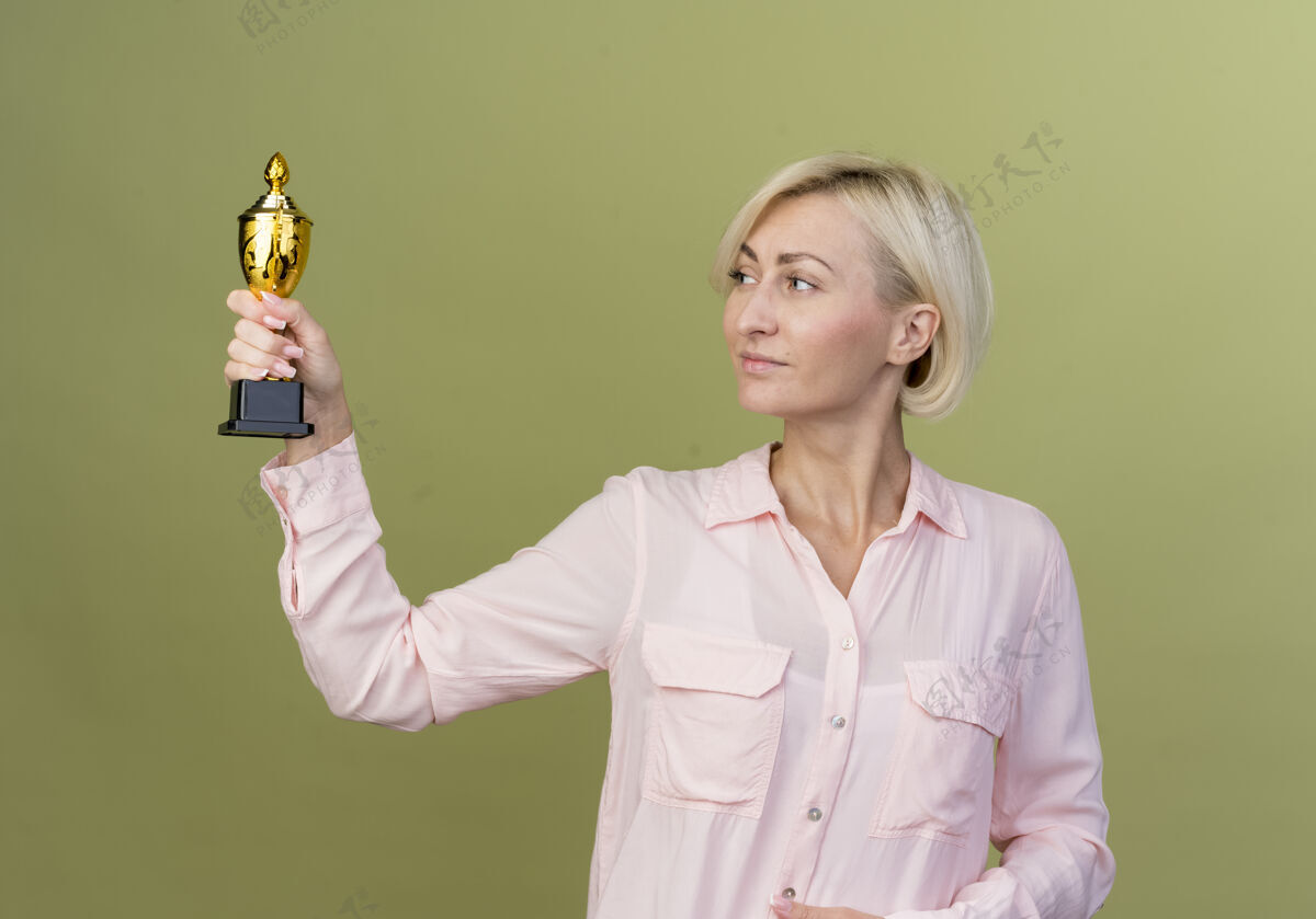 金发高兴的年轻金发斯拉夫女子拿着冠军杯看绿色优胜者女士