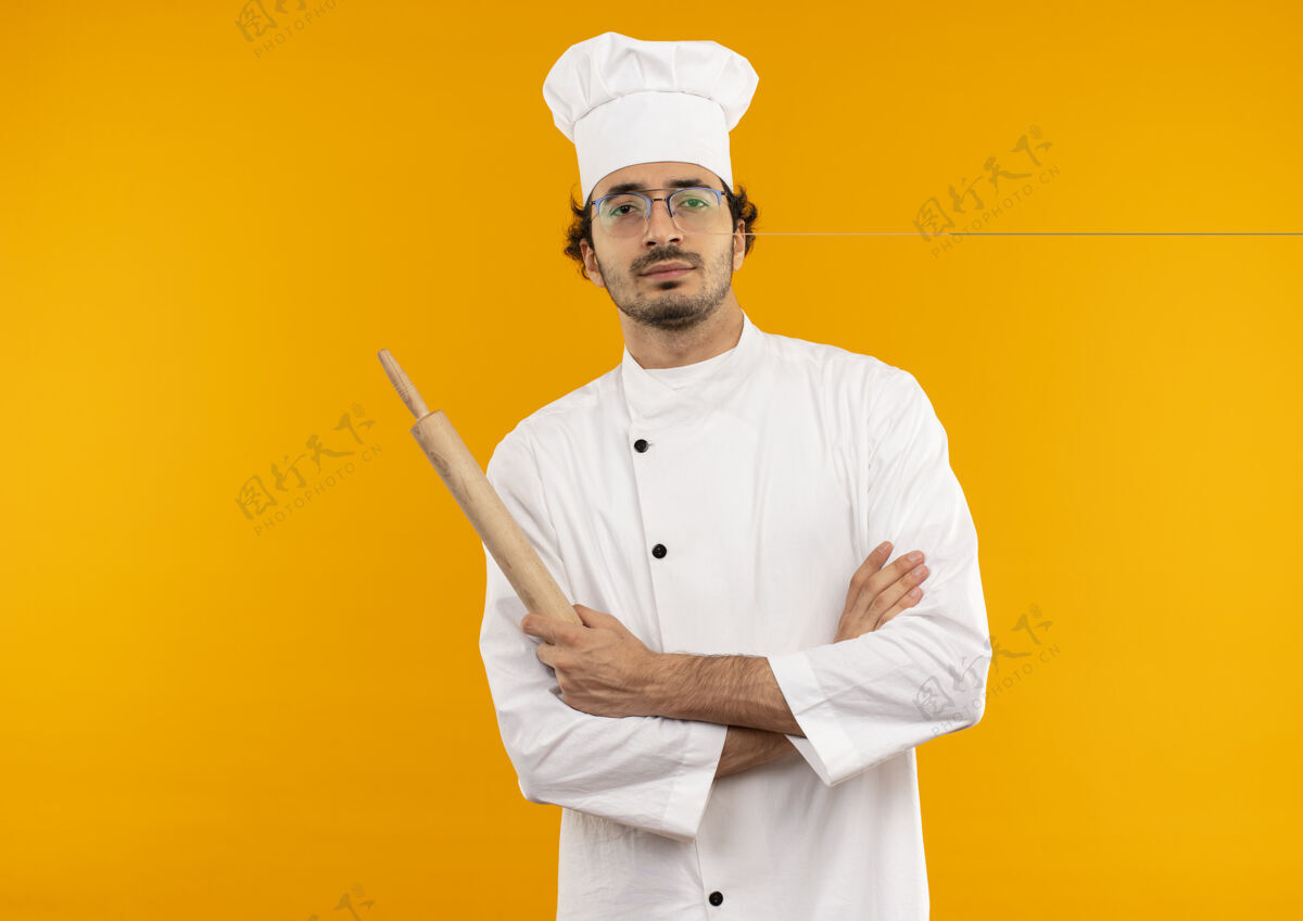 男人自信的年轻男厨师穿着厨师制服 戴着眼镜 双手交叉 手持擀面杖年轻穿男性