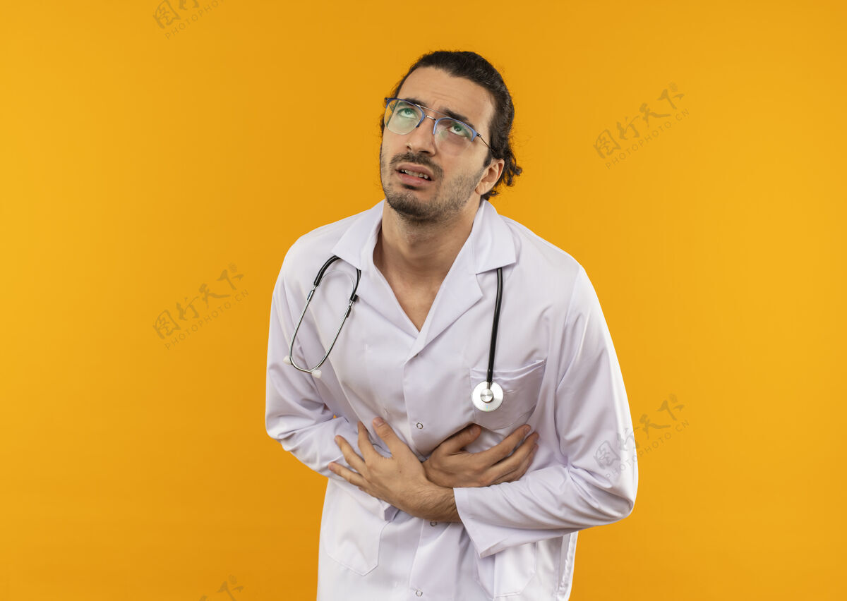听诊器看着生病的年轻医生戴着医用眼镜穿着医用长袍用听诊器抓住了肚子男人医生医疗
