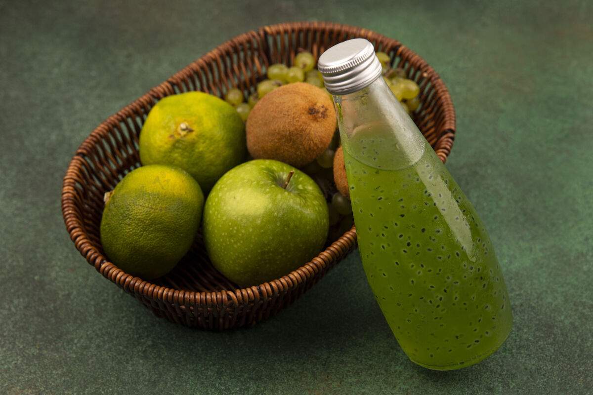 食物侧视图绿色的橘子与苹果猕猴桃和葡萄在一个篮子与一瓶果汁在绿色的墙上苹果瓶子景观