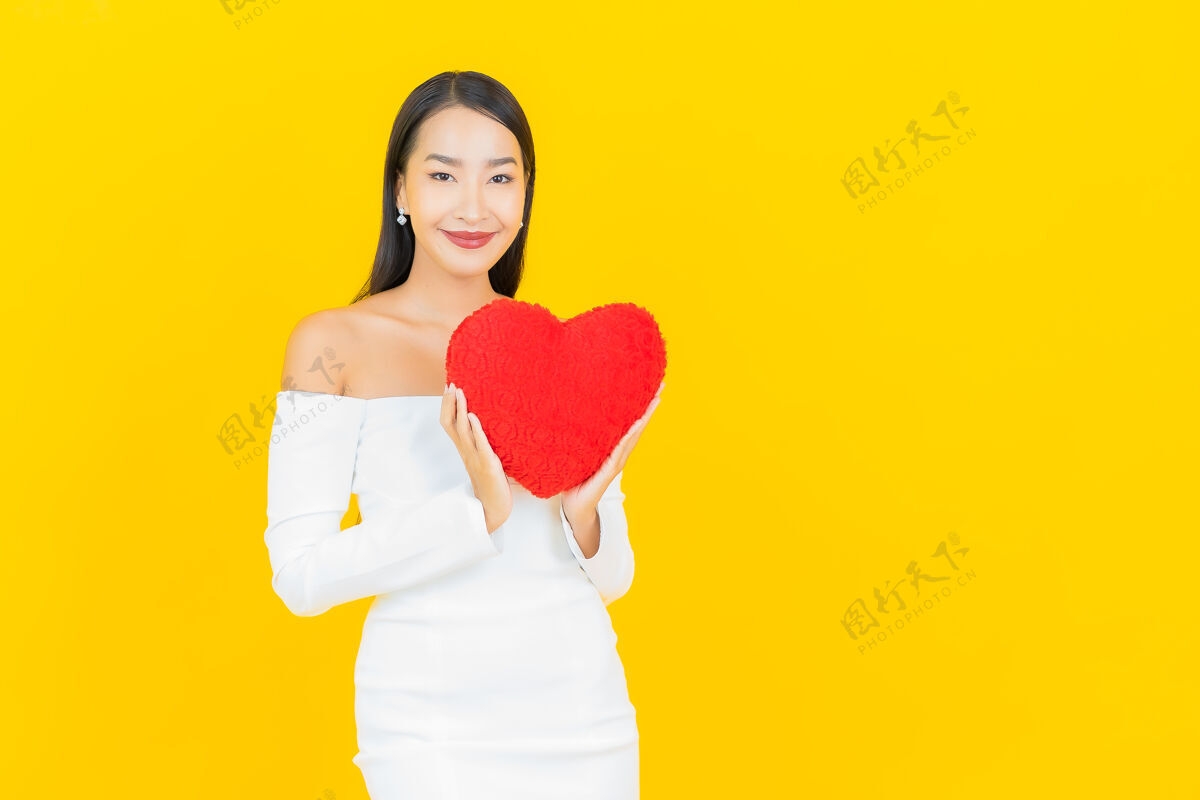 美丽黄色墙壁上有一个心形枕头的亚洲商界年轻美女的画像情人节年轻可爱